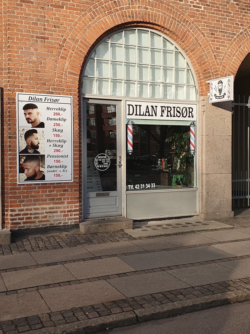 Butikken på Finsensvej 52: røde mursten, rundbue, hvidt skilt med Dilan Frisør og prisskiltet på muren.